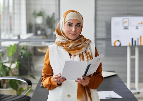 A professional Muslim woman in an office setting, confidently holding documents and smiling.