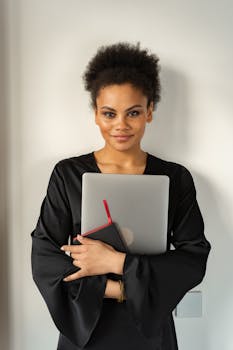 Smiling woman with curly hair holding a laptop and notebook indoors.