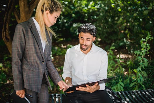 Two business professionals discussing documents on a sunny park bench.