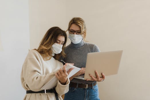 Two women in masks working indoors with a laptop and notebook, highlighting remote work during the pandemic.